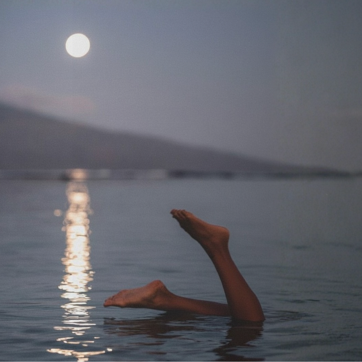 A person's bare legs and feet extend out of calm water under a full moon, with a soft mountain silhouette in the background.